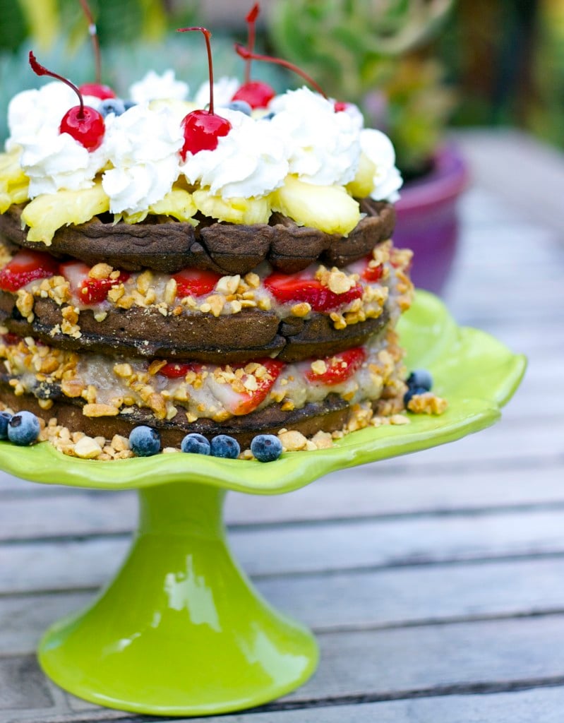 Banana split waffle cake with alternating layers of chocolate Belgian waffle and strawberries with peanuts, topped with whipped cream and maraschino cherries on a lime green cake stand. 