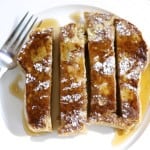 One slice of french toast drizzled in maple syrup, garnished with powdered sugar and cut in to four strips on a white plate from bird's eye view.
