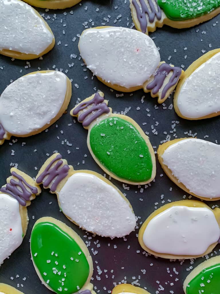 Overhead photo of sugar cookies shaped like christmas tree bulbs, iced with green or white icing and sprinkled with coarse sugar on black tray