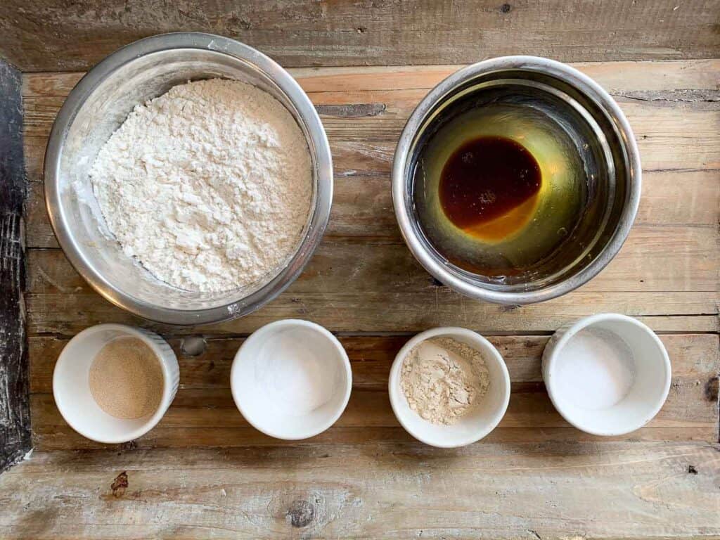 Overhead view of bagel ingredients in bowls on wooden surface (flour, water/oil/barley malt syrup; instant yeast; salt; vital wheat gluten; sugar