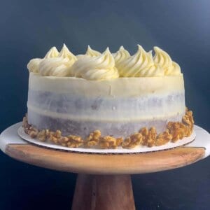 side view of carrot cake with cream cheese frosting, including rosettes, shredded carrot and walnuts, on marble and wood cake stand with black background.
