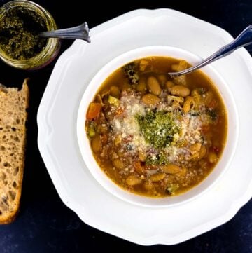 overhead view of minestrone soup in white bowl with pesto and parmesan topping, slice of sourdough bread, and jar of pesto, all on black background.