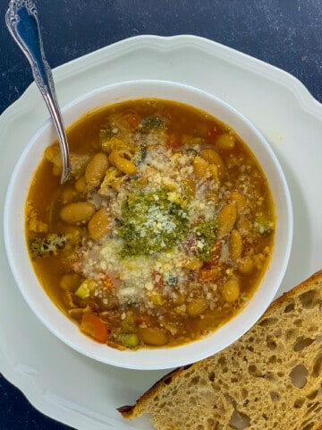 top view of minestrone in white bowl with bread grated cheese and spoon