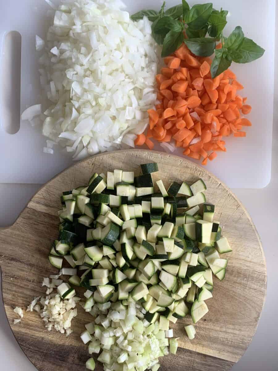 chopped garlic, celery, zucchini on wooden chopping board; chopped onion, carrot and spring of basil on white chopping board How To Make the Best Homemade Minestrone
