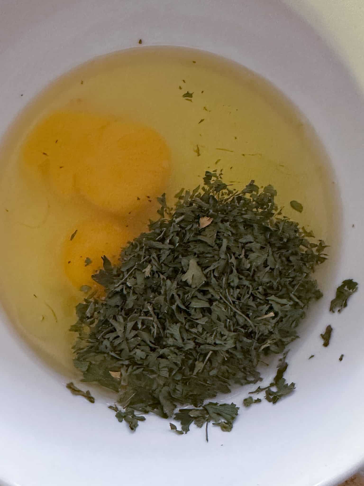 close up view of dried parsley, egg, and olive oil in white bowl