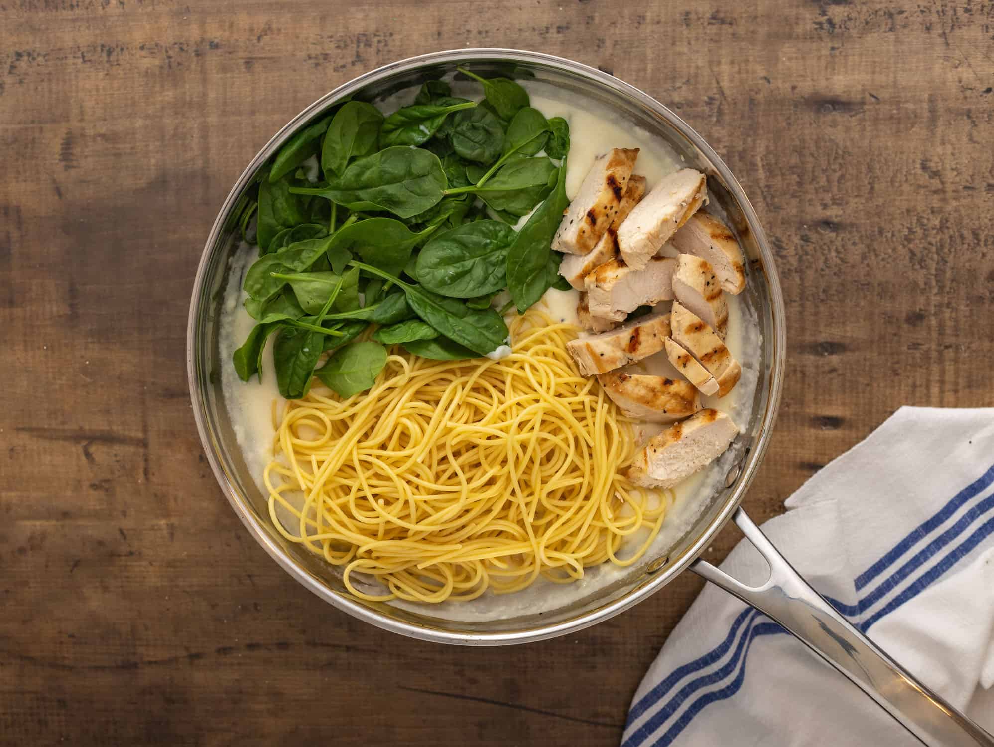 Adding pasta, spinach and chicken to cream sauce in pan.