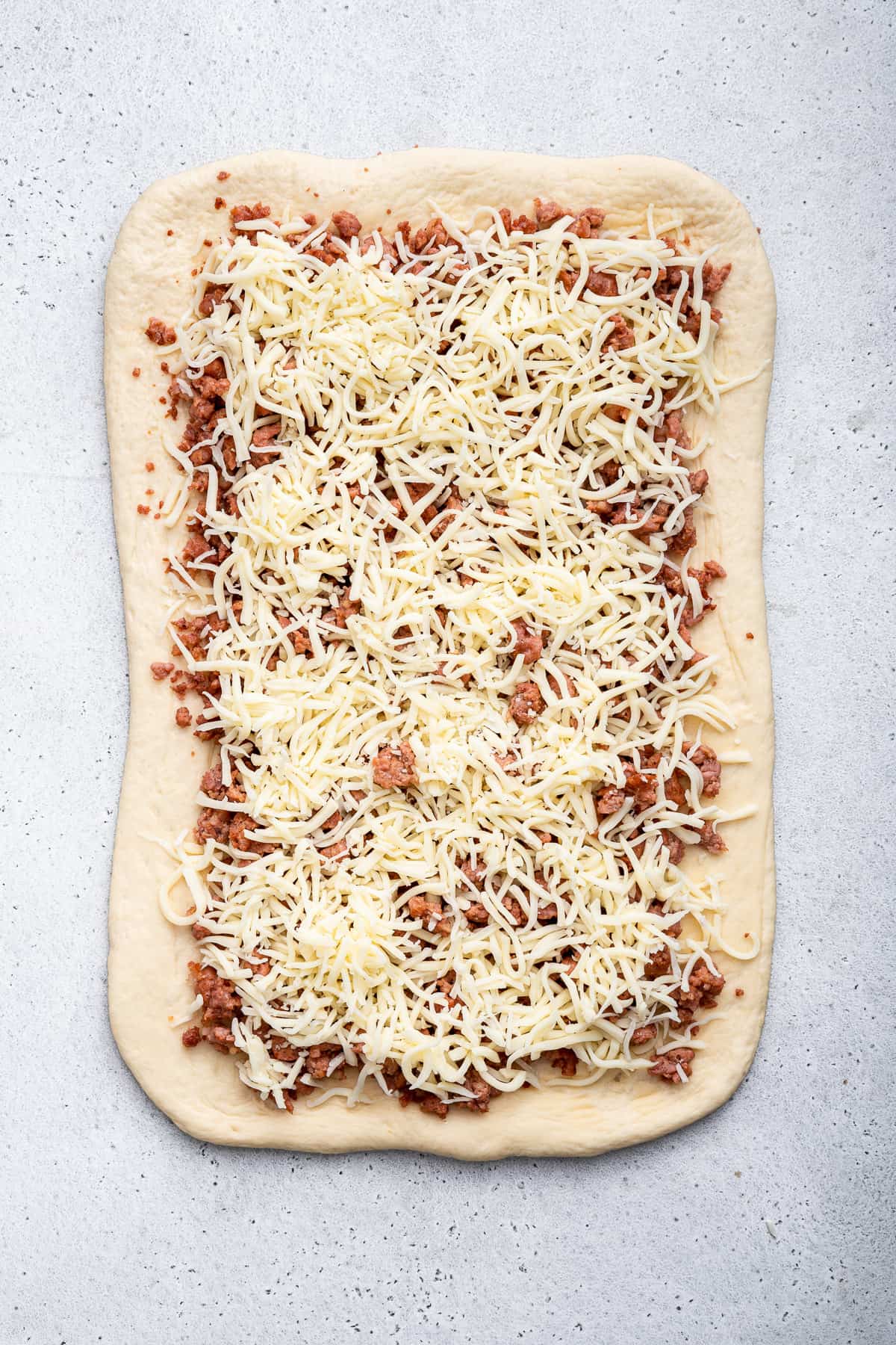 Rectangle of dough topped with cooked sausage and shredded cheese.