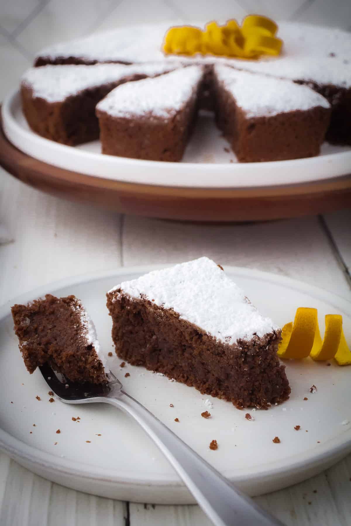 Slice of torte caprese on a plate with a fork and cake in the background.