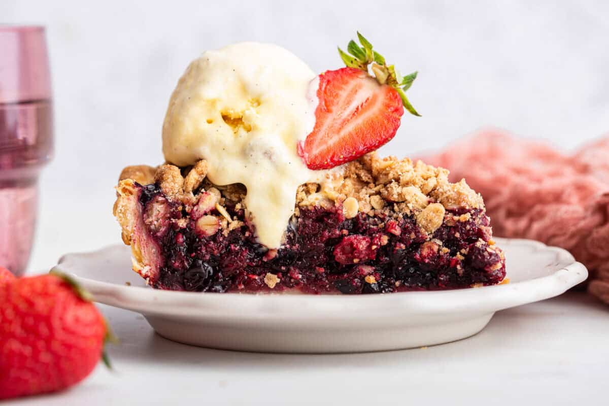 Close up of a berry pie slice on white plate topping with vanilla ice cream and a sliced strawberry.