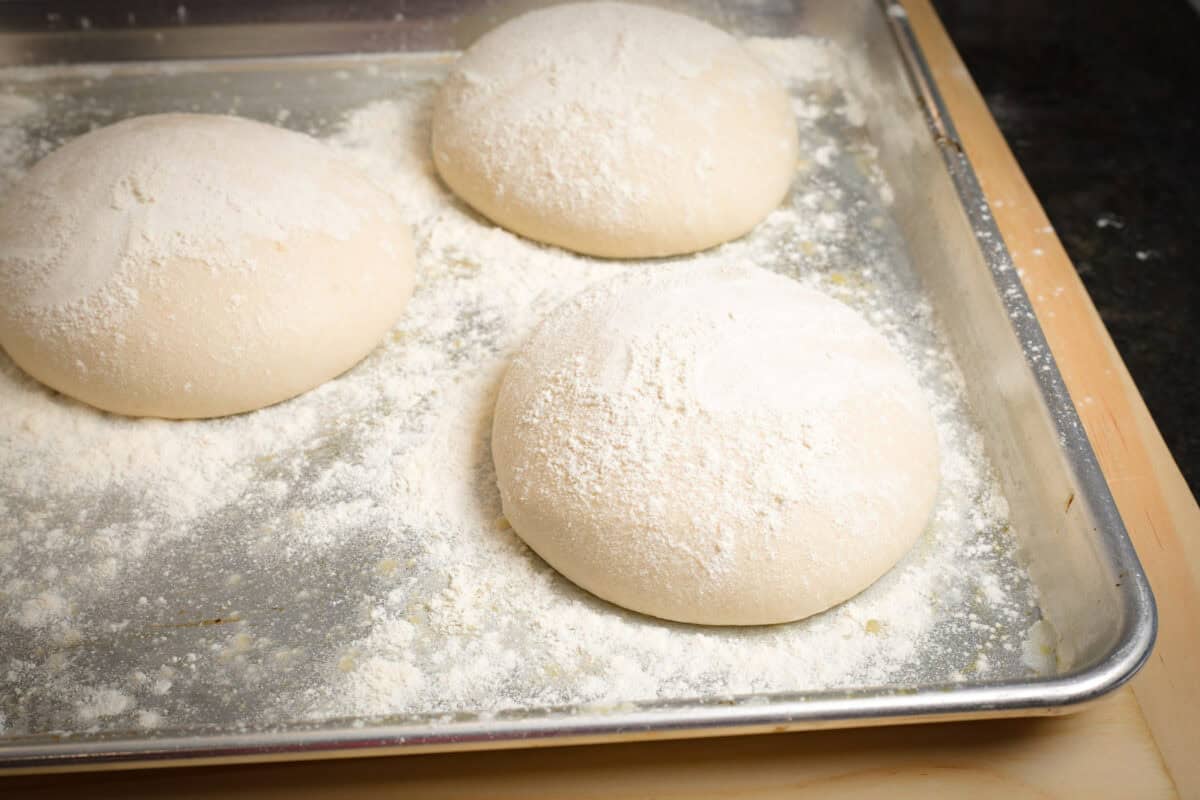 Three pizza dough balls dusted with flour on baking sheet.