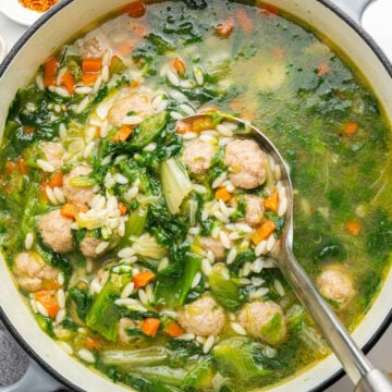 White pot with spoon full of Italian wedding soup with escarole, mini meatballs, carrots and small pasta.