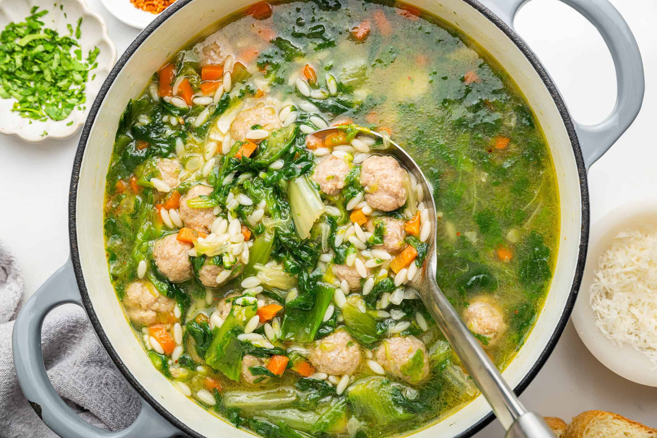 White pot with spoon full of Italian wedding soup with escarole, mini meatballs, carrots and small pasta. 