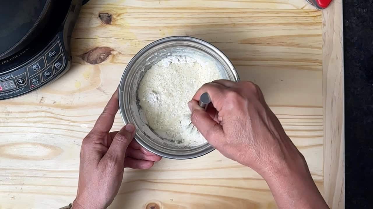 A person stirs a white batter in a metal bowl with a spoon on a light wooden surface. An induction cooktop is partially visible on the left side.