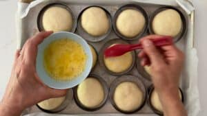 A person brushes egg wash onto round dough balls in baking rings on a tray, preparing homemade hamburger buns for baking. The left hand holds a bowl of whisked eggs while the right hand uses a red pastry brush.