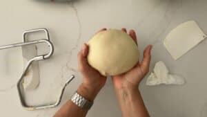 A person holding a smooth ball of dough for homemade hamburger buns over a marble countertop, with a dough hook, dough scraper, and a piece of paper towel nearby.