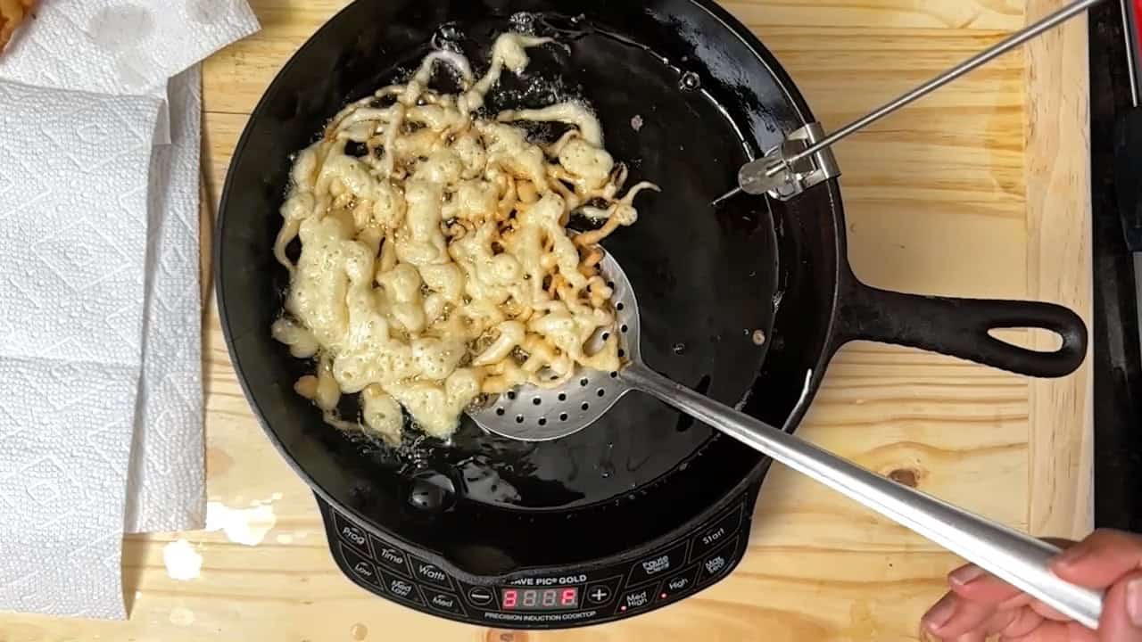 A person uses a slotted spatula to fry and lift funnel cake batter in hot oil on a black stovetop, with paper towels nearby on a wooden counter.