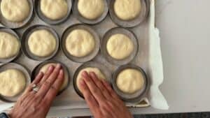 Hands press dough into round rings on a parchment-lined baking sheet, preparing evenly spaced homemade hamburger buns for baking.