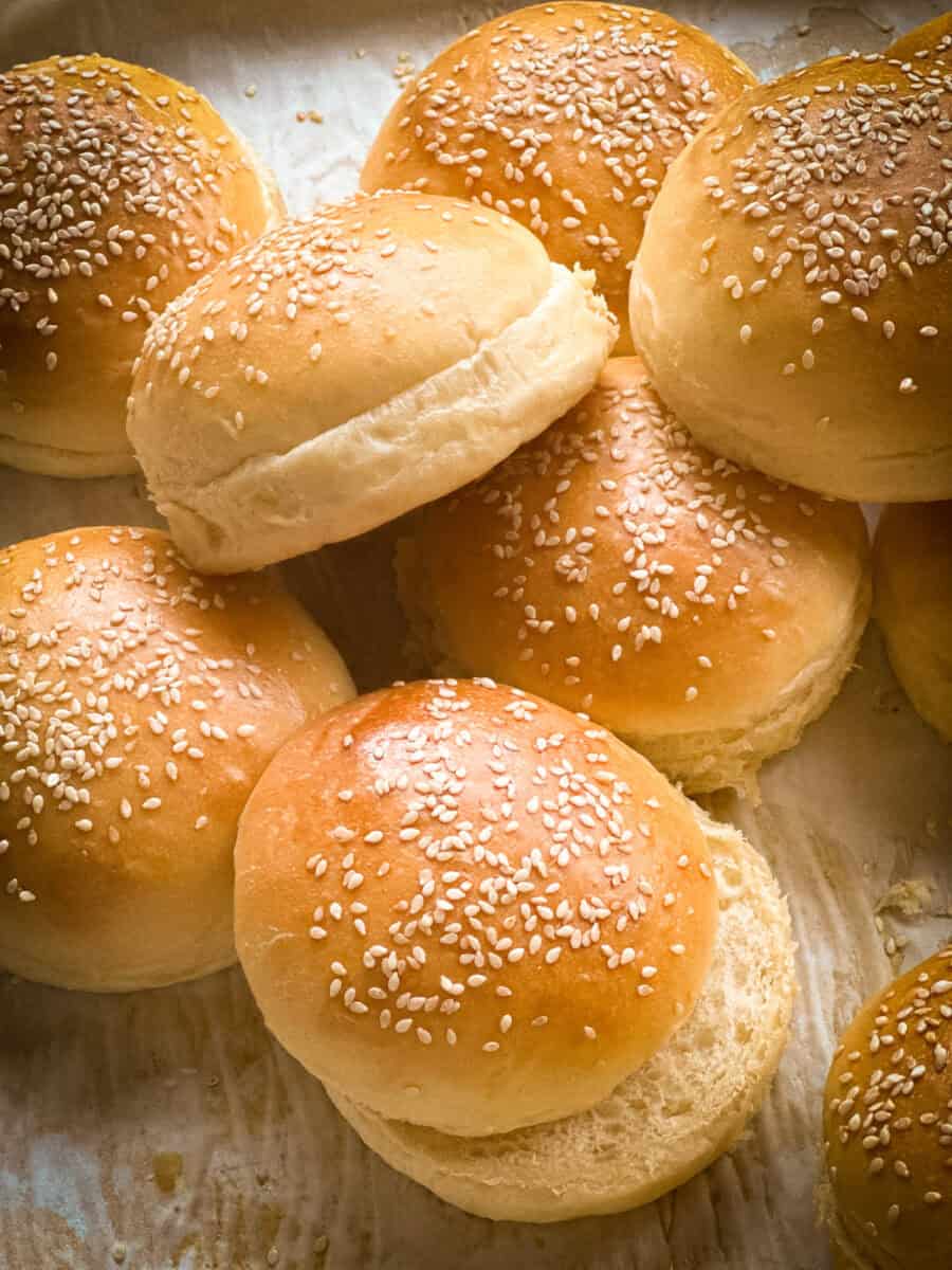 A close-up of several golden brown hamburger buns topped with sesame seeds, arranged on a light-colored surface. Some buns are slightly overlapping, and one is split open to show the soft inside.