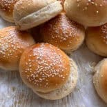 A close-up of several golden brown sesame seed hamburger buns on parchment paper, some stacked and some with tops slightly askew, showing their soft, fluffy texture.