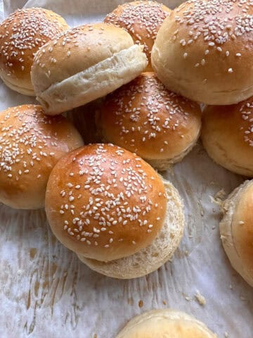 A close-up of several golden brown sesame seed hamburger buns on parchment paper, some stacked and some with tops slightly askew, showing their soft, fluffy texture.