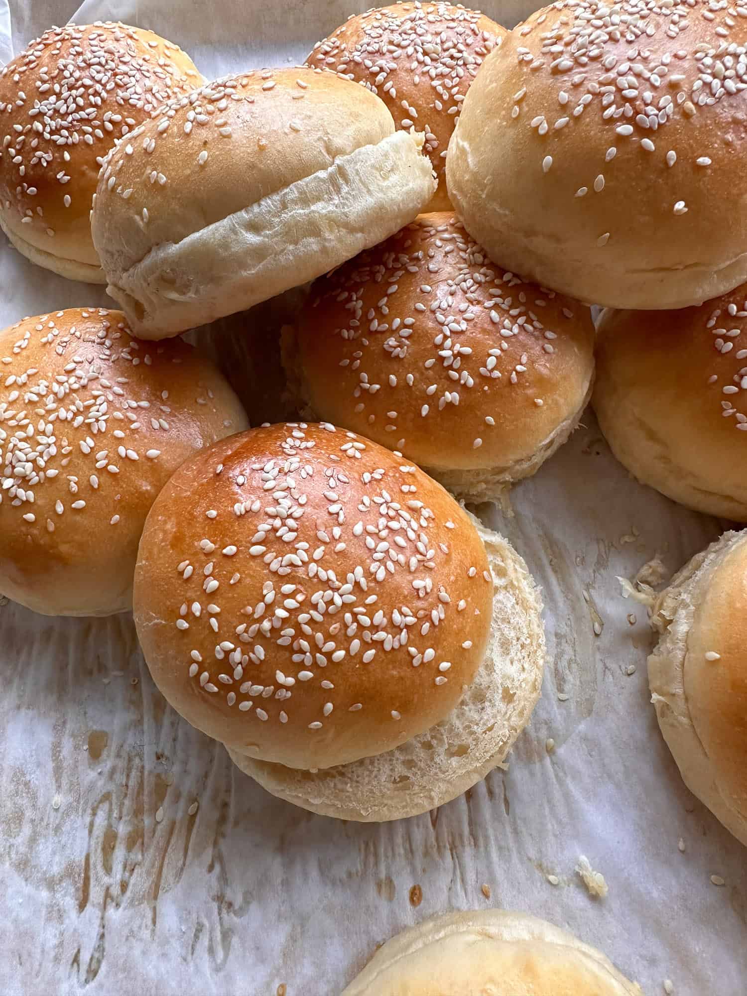 A close-up of several golden brown sesame seed hamburger buns on parchment paper, some stacked and some with tops slightly askew, showing their soft, fluffy texture.