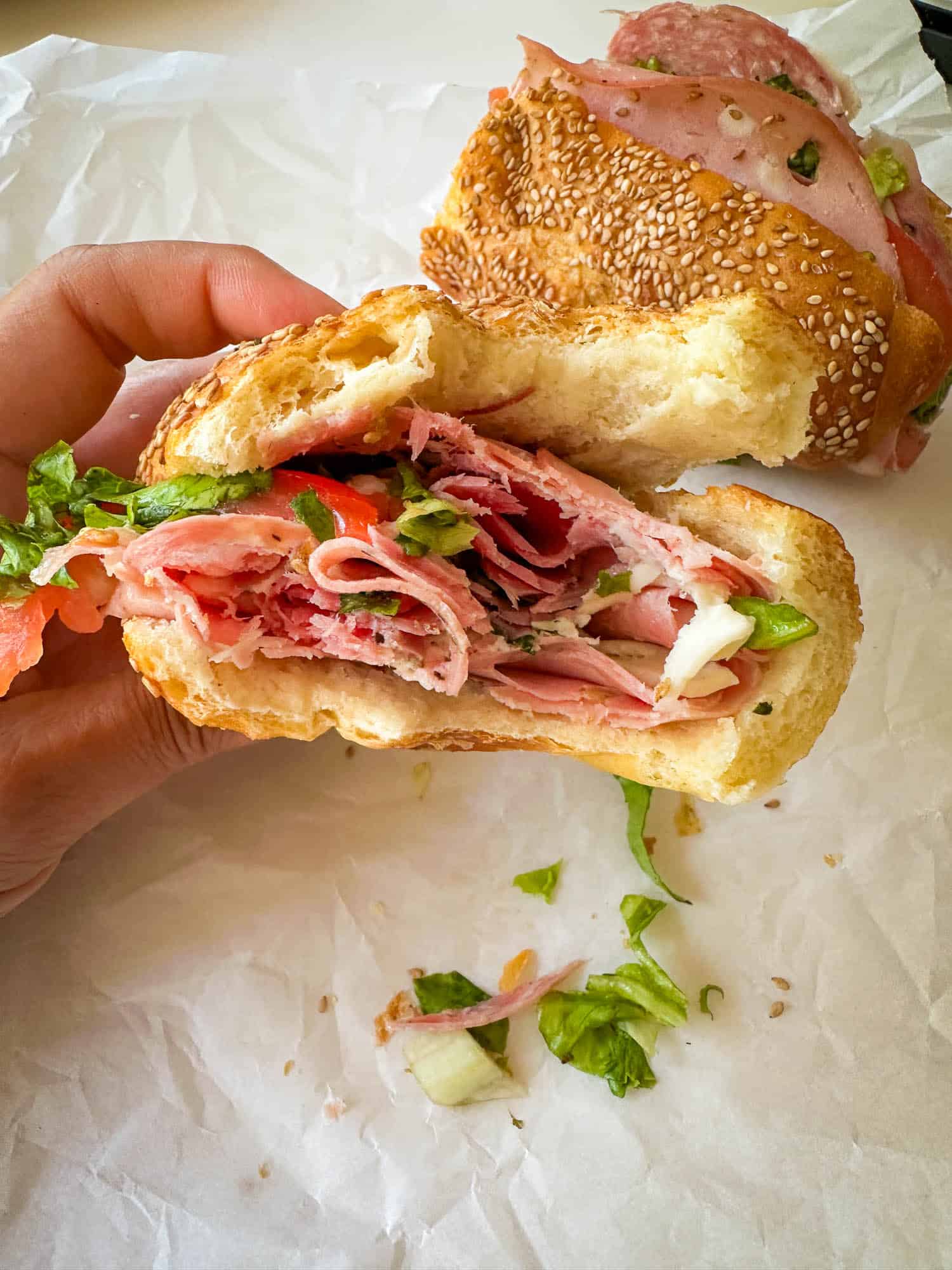 A hand holds a half-eaten sandwich filled with layers of deli meats, lettuce, and other toppings on a sesame seed bun. Another sandwich half rests in the background on white parchment paper.