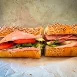 A sandwich on a sesame seed hoagie roll, cut in half, filled with layers of deli meat, lettuce, tomato, and cheese, sitting on parchment paper against a neutral background.