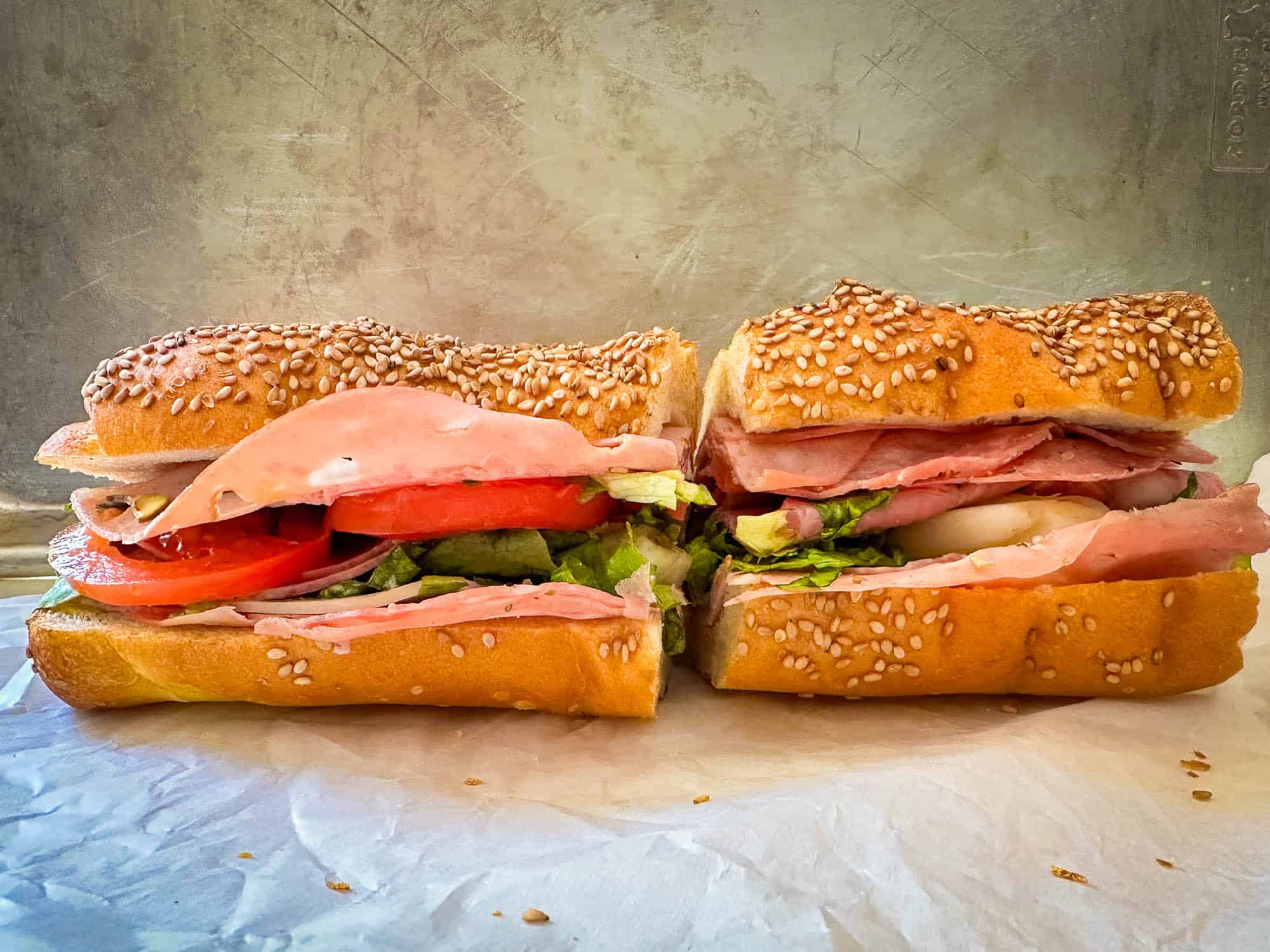 A sandwich on a sesame seed hoagie roll, cut in half, filled with layers of deli meat, lettuce, tomato, and cheese, sitting on parchment paper against a neutral background.
