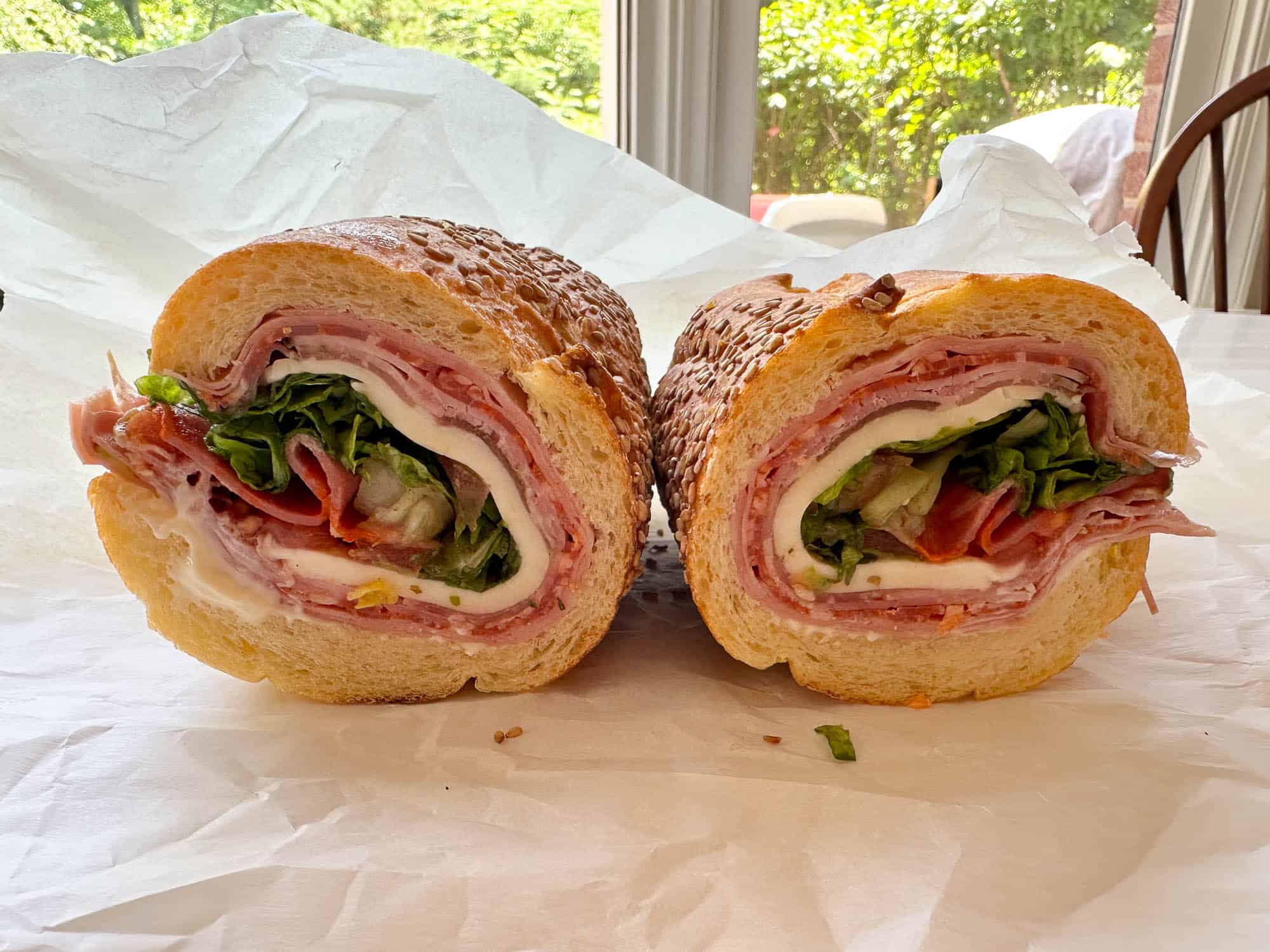 A large Italian sub sandwich cut in half, showing layers of deli meats, cheese, lettuce, tomato, and onions inside a sesame seed hoagie roll, placed on white sandwich paper with a bright window and greenery in the background.
