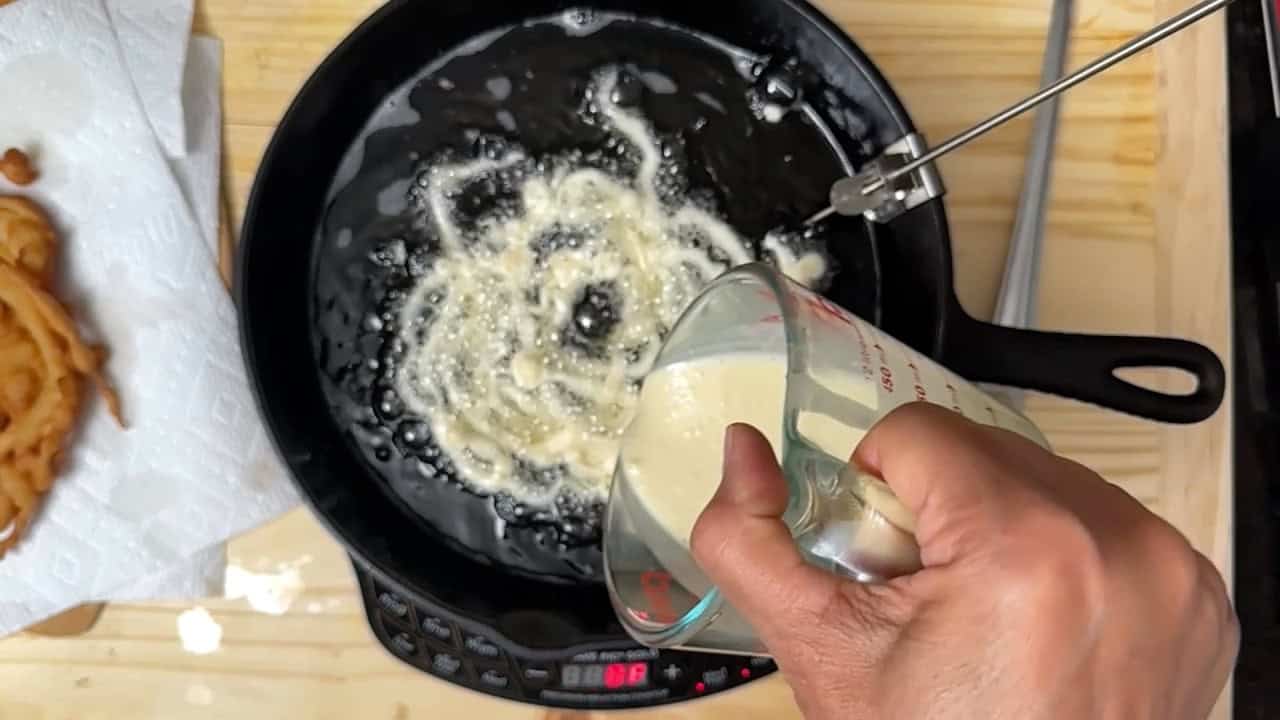 A hand pours batter from a measuring cup into a black skillet filled with hot oil on a stovetop, with bubbles forming. Fried pieces rest on a paper towel nearby.