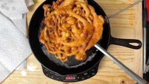 A funnel cake is being fried in a black cast iron skillet on an electric cooktop, with metal tongs holding the edge. Paper towels are set nearby on a wooden countertop.