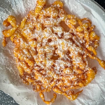 A classic funnel cake topped with powdered sugar sits on parchment paper atop a white plate, with extra sugar scattered on the black surface below—capturing the irresistible charm of homemade funnel cakes.