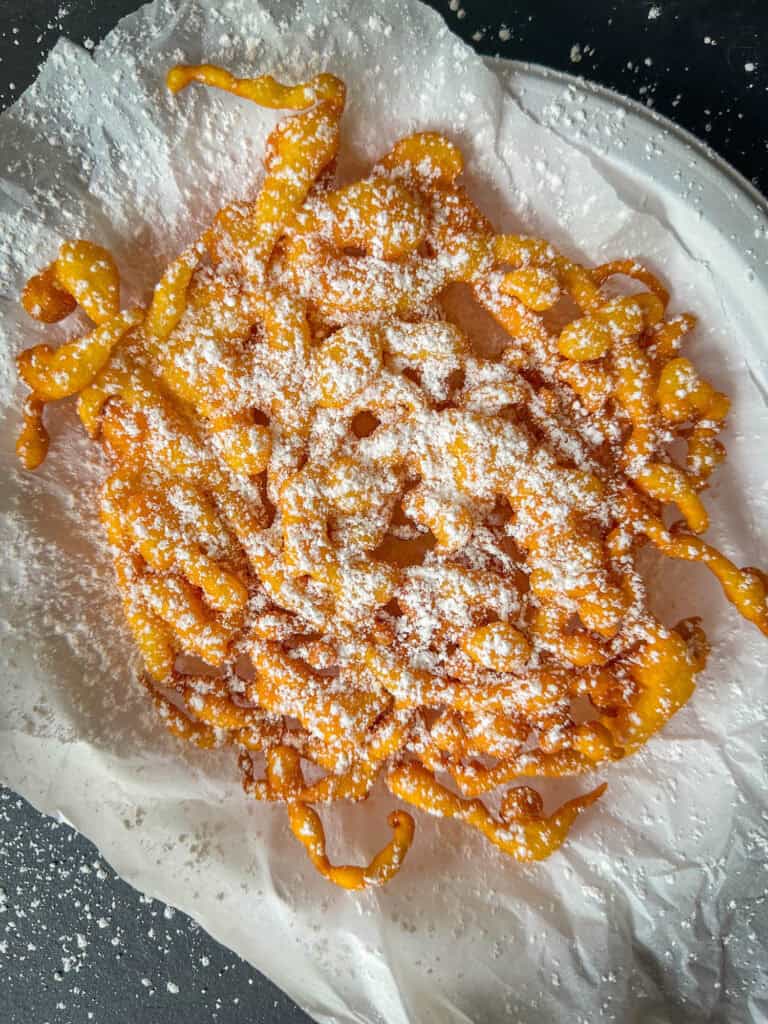 A classic funnel cake topped with powdered sugar sits on parchment paper atop a white plate, with extra sugar scattered on the black surface below—capturing the irresistible charm of homemade funnel cakes.