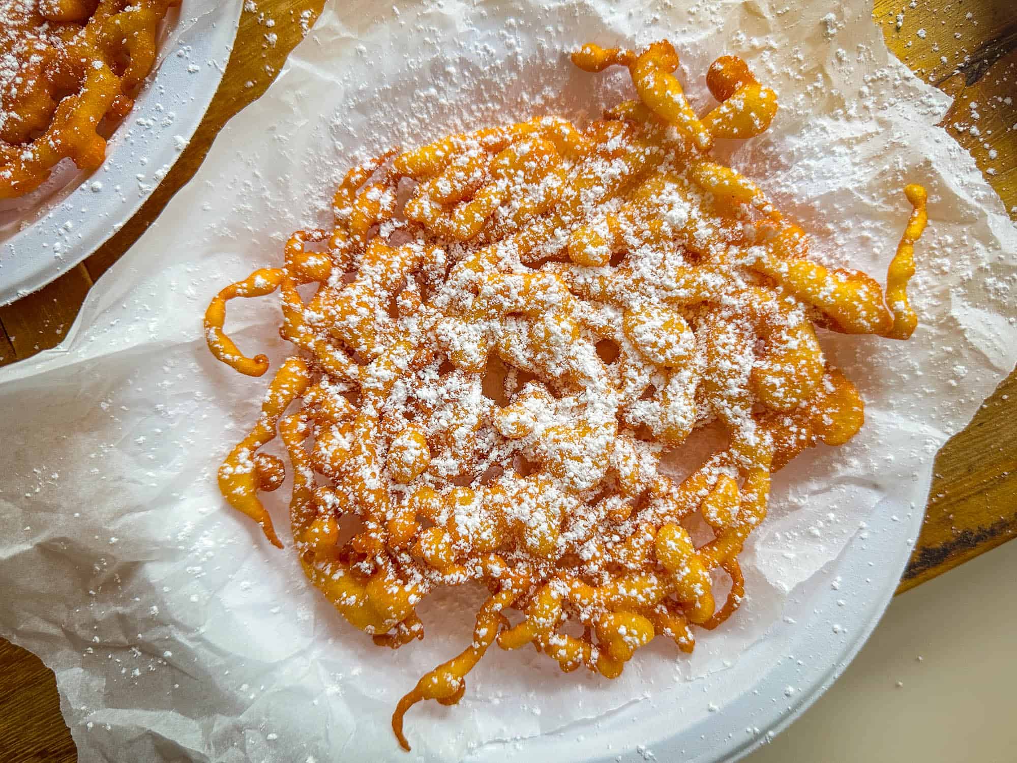 A plate of classic funnel cakes dusted with powdered sugar, served on parchment paper atop a wooden surface.