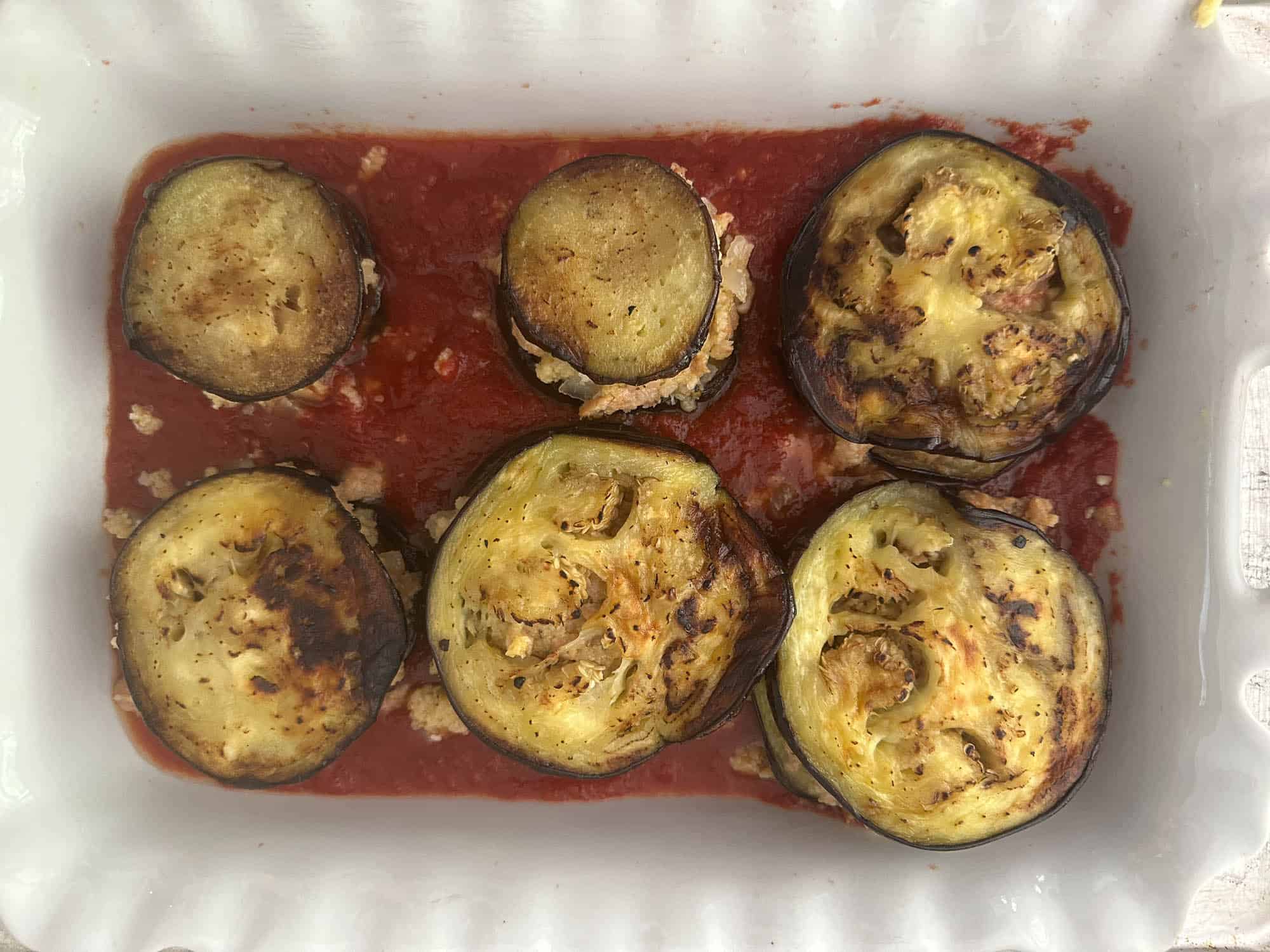 Roasted eggplant slices stuffed with sausage bread stuffing in a tomato sauce on a white baking dish.